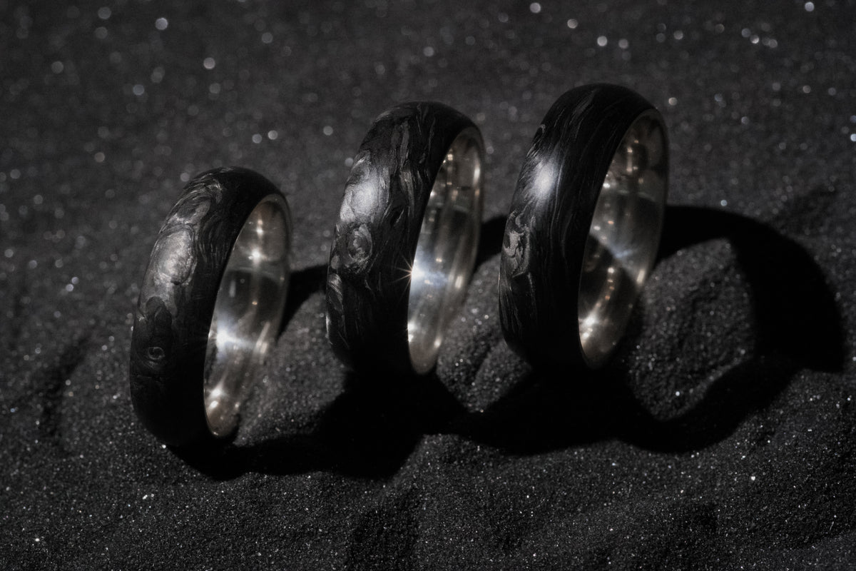 Three sterling silver and forged carbon fiber mens wedding bands on black sand