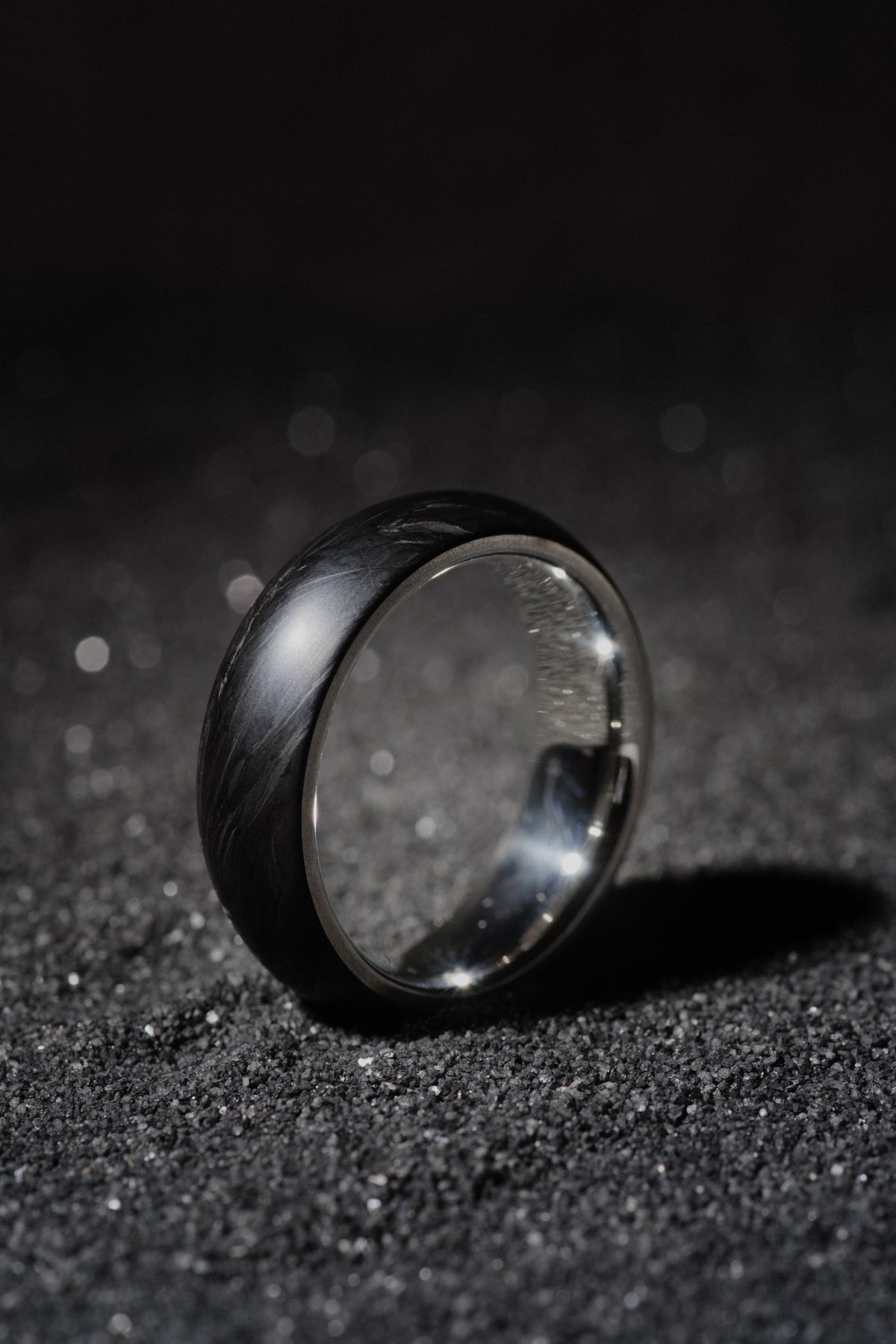 Sterling silver and forged carbon fiber mens wedding band standing on black sand